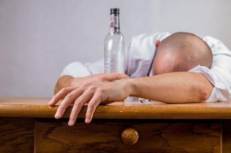 Ein Mann liegt mit dem Oberkörper auf einem Tisch, eine Alkoholflasche wird von einem Hand gegeriffen während die andere lässig davor liegt.