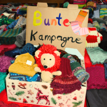 Ein kleiner gestrickter Löwe guckt aus einer Schachtel. Darüber steht gechrieben: "Bunte Socken Kampagne". Im Hintergrund liegen viele Socken.